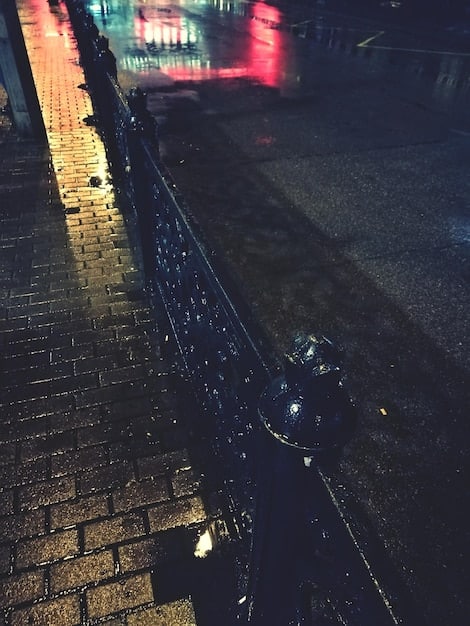 A scene from Dark City showing John Murdoch (Rufus Sewell) standing in the rain-soaked, dimly lit streets of the city. The architecture should appear distorted and unsettling.