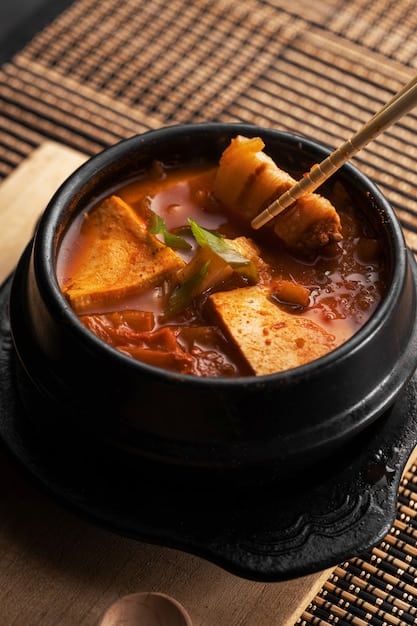 A close-up shot of a steaming bowl of Kimchi Jjigae (Kimchi Stew) with visible tofu, pork, and kimchi, garnished with green onions. The photo should highlight the rich, red color and the bubbling texture of the stew, conveying a sense of warmth and spiciness.