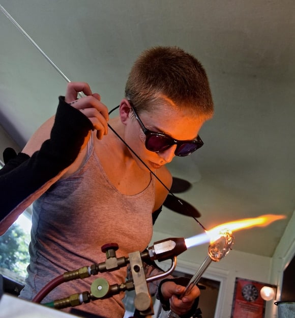 A person wearing safety goggles working on soldering wires inside the hilt of a lightsaber. The workbench is organized with various electronic components and tools surrounding them. The focus is on the meticulous detail and precision required for electronics work in a DIY setting.
