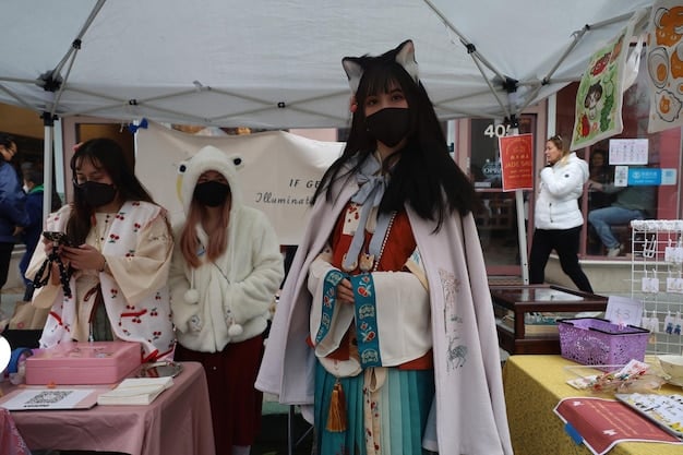 A wide shot of a cosplay vendor booth at a bustling convention. Several pre-made costumes are displayed on mannequins, showing various characters from anime, video games, and comics. Cosplayers are browsing the selection, and the booth is filled with colorful props and accessories.