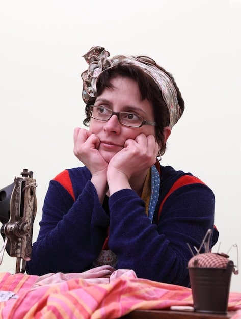A cosplayer working at a table covered in fabric scraps, patterns, and sewing tools, carefully stitching a piece of fabric for their costume, with a focused expression.