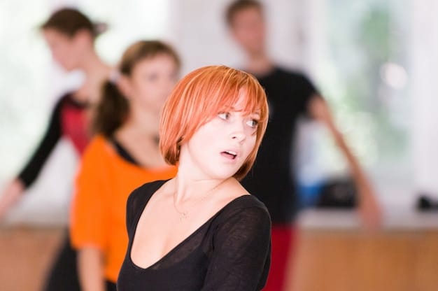A close-up of a K-Pop choreographer teaching a group of dancers a complex move. The choreographer, dressed in street style, is demonstrating sharp, precise movements. The dancers are focused and mirroring the choreographer's actions, reflecting the detailed and rigorous training involved in K-Pop dance.