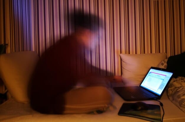 An individual sitting alone in a dimly lit room, looking at a laptop with a worried expression. The lighting and body language convey feelings of isolation, anxiety, and distress.