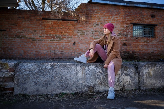 A street style shot featuring a young woman wearing an oversized pastel pink hoodie, ripped jeans, chunky white sneakers, and a baseball cap. She is posing confidently on a city street.