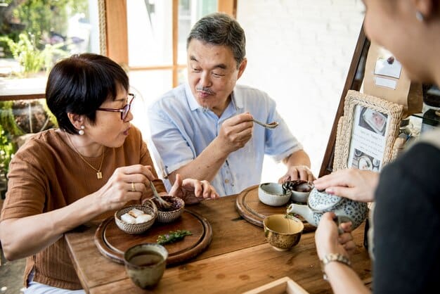 Three participants, two women and one man, seated in a traditional Korean tea room, participating in a tea ceremony. The women are wearing elegantly simple, modern hanboks in muted, earthy tones, while the man is wearing a respectful business casual outfit. They are all seated on cushions around a low tea table set with traditional celadon teaware. The room has traditional Korean paper windows, allowing soft, diffused light to fill the space, highlighting the tranquility and simplicity of the ceremony.]