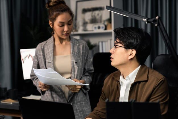 Two Korean business professionals in a meeting room, discussing documents. One is pointing to a chart while the other listens attentively. The setting is a bright and modern office.