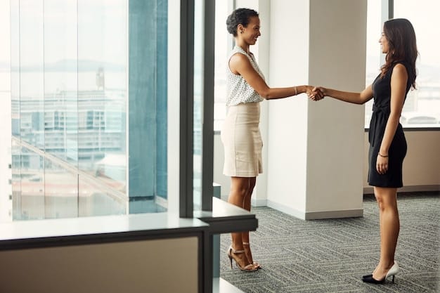 A US professional shaking hands with a Korean colleague in a modern office setting. Both are smiling and appear to be engaged in a friendly conversation. The background includes other colleagues working at their desks.