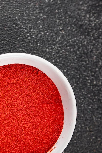 A close-up shot of gochugaru (Korean chili flakes) in a small bowl, highlighting its vibrant red color and coarse texture. A hand sprinkles the gochugaru onto a pile of napa cabbage in a larger bowl.