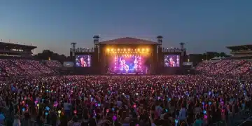 Thousands of K-pop fans at a lively outdoor music festival in the US, cheering with lightsticks as a K-pop idol performs on a brightly lit stage.