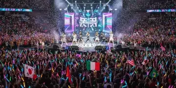 K-Pop group performing live on a brightly lit stage in a packed arena, surrounded by cheering fans with lightsticks.